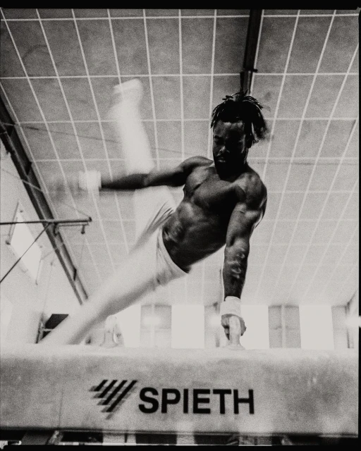 Black and white photo of a male gymnast performing on a pommel horse in a gymnasium with overhead lighting and athletic equipment.