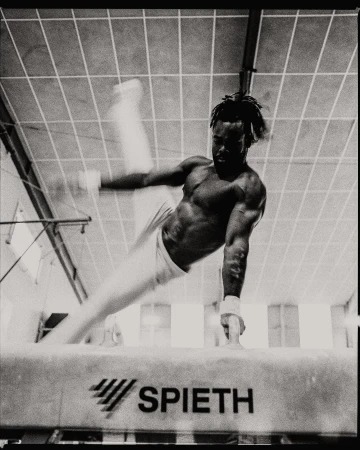 Black and white photo of a male gymnast performing on a pommel horse in a gymnasium with overhead lighting and athletic equipment.