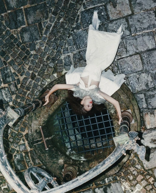 Woman in an elaborate white gown lies near a weathered stone well, shot from a high angle showcasing a blend of fashion and historical architecture.