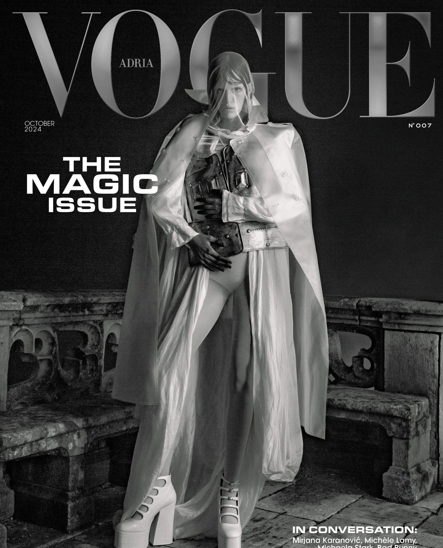 Vogue cover in black and white featuring a model in a white hooded cape, corset, shorts, and platform heels standing on a stone balcony.