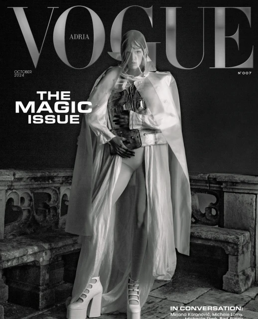 Vogue cover in black and white featuring a model in a white hooded cape, corset, shorts, and platform heels standing on a stone balcony.
