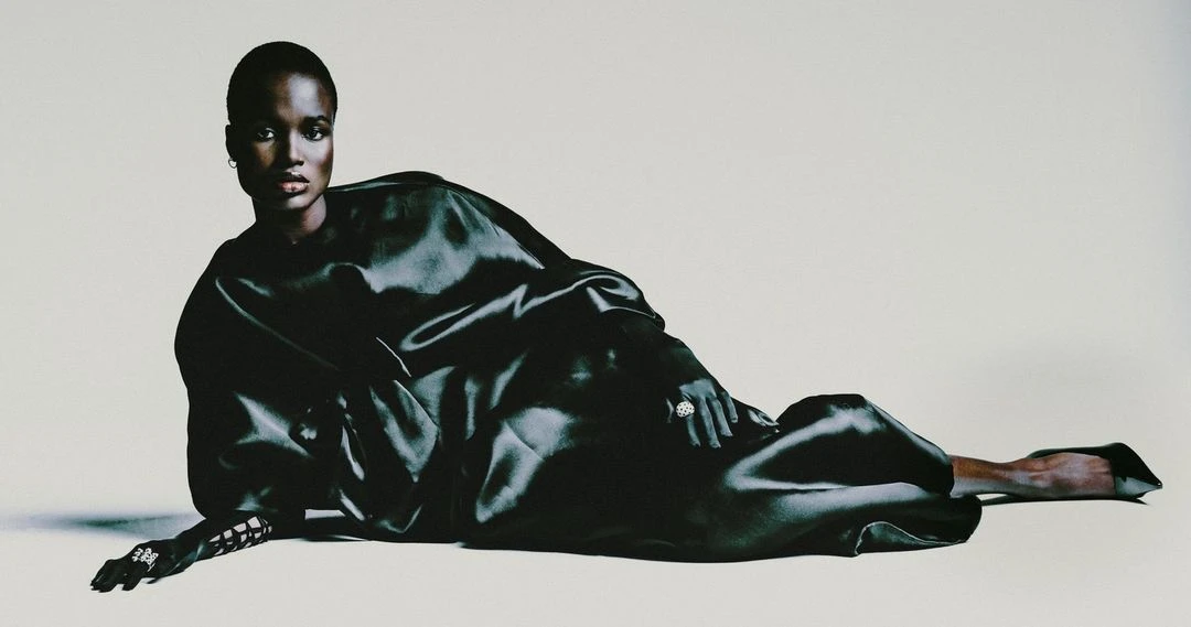 Elegant African American woman in a black satin dress reclines against a white backdrop, showcasing fashion and style with dark tones.