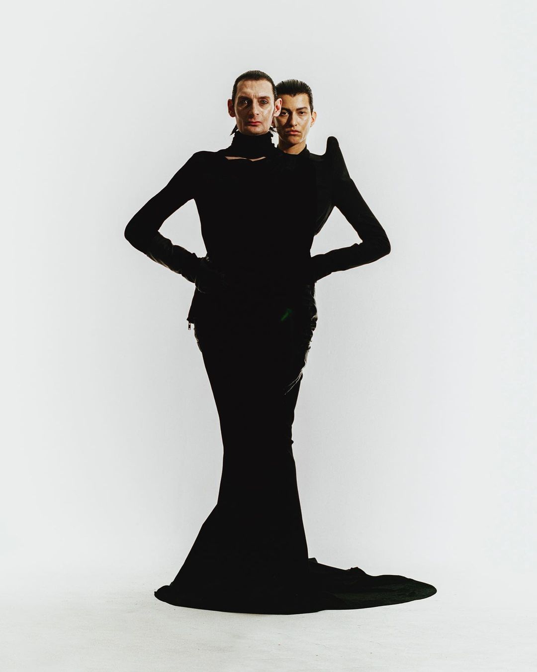 Two fashion models in black outfits pose in a photo studio against a white background. The first model stands in front of the other.