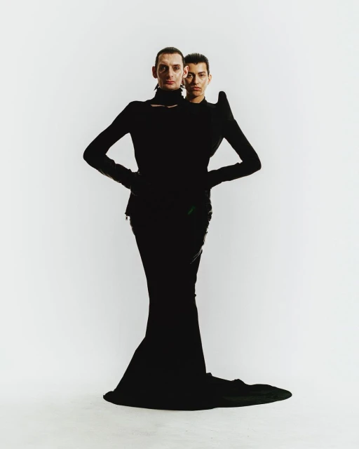 Two fashion models in black outfits pose in a photo studio against a white background. The first model stands in front of the other.