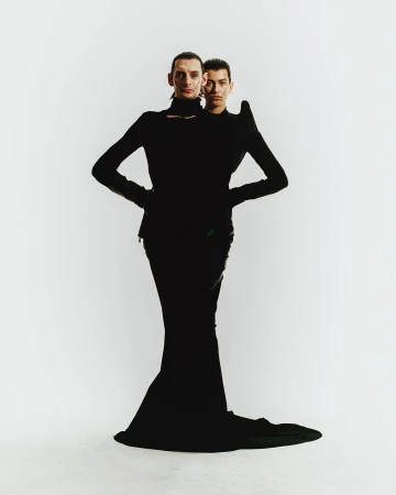 Two fashion models in black outfits pose in a photo studio against a white background. The first model stands in front of the other.