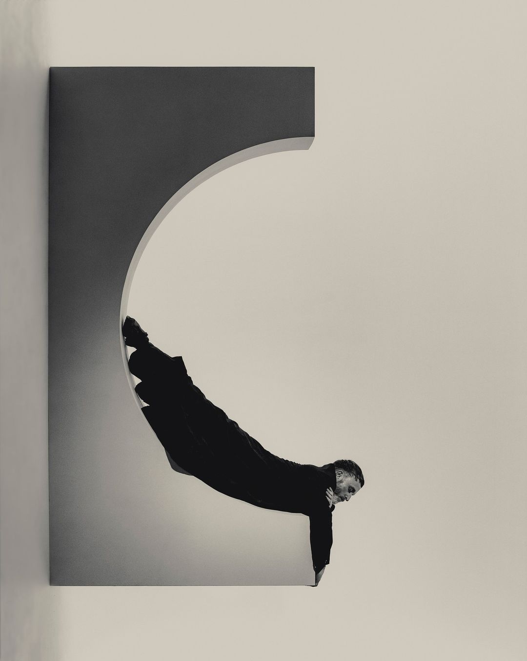 Adult male in black clothing performing a physical feat within a rectangular architectural opening with an arched section, captured in a studio.