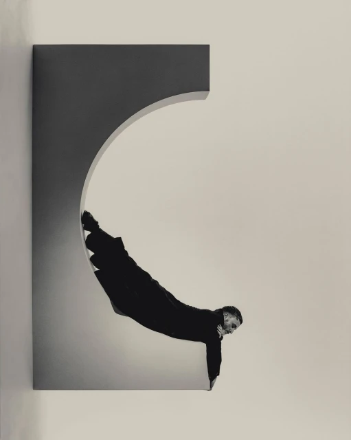 Adult male in black clothing performing a physical feat within a rectangular architectural opening with an arched section, captured in a studio.