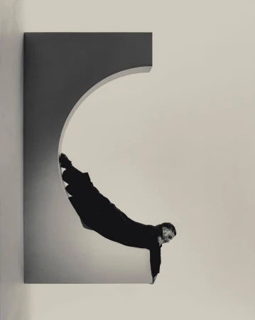 Adult male in black clothing performing a physical feat within a rectangular architectural opening with an arched section, captured in a studio.