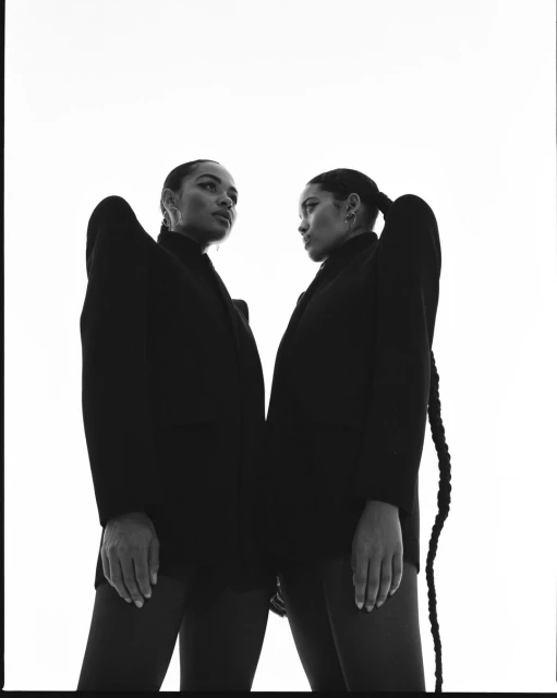 Two women pose in black blazers against a bright white backdrop, creating a striking contrast in a high-fashion studio portrait.