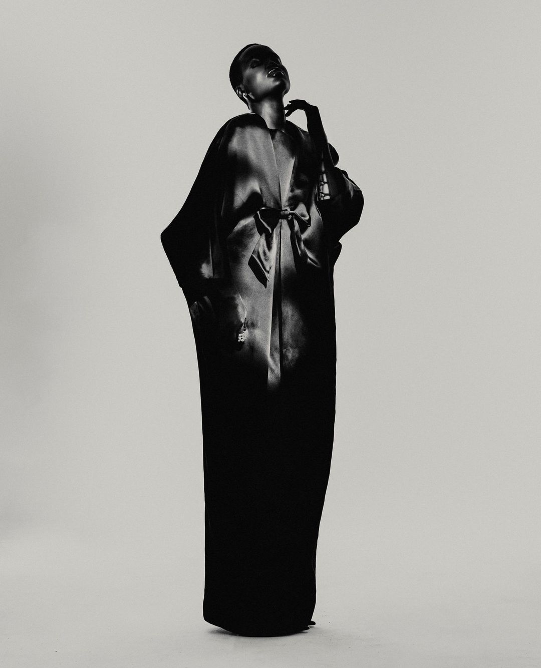 Black and white fashion shot of a woman in long robe, posed against a light background, emphasizing texture and form in a studio setting.