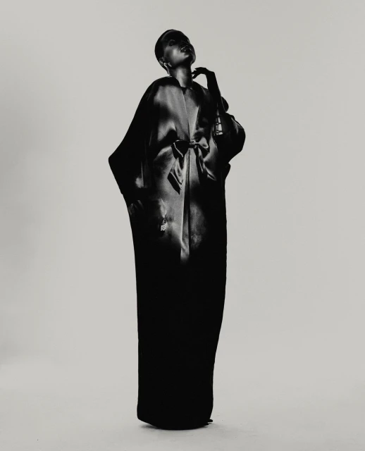 Black and white fashion shot of a woman in long robe, posed against a light background, emphasizing texture and form in a studio setting.