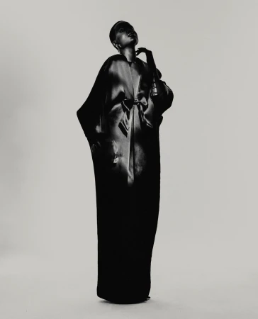 Black and white fashion shot of a woman in long robe, posed against a light background, emphasizing texture and form in a studio setting.