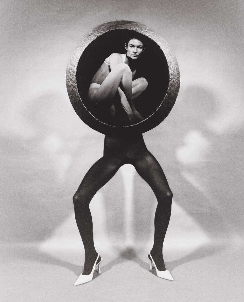 Black-and-white fashion shot featuring a woman in a bowl on the legs of a figure in tights and heels against a white background.