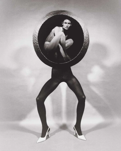 Black-and-white fashion shot featuring a woman in a bowl on the legs of a figure in tights and heels against a white background.