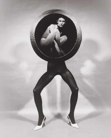 Black-and-white fashion shot featuring a woman in a bowl on the legs of a figure in tights and heels against a white background.