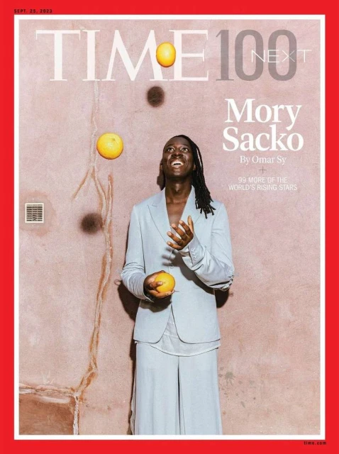 Magazine cover featuring Mory Sacko juggling oranges, wearing a light blue suit against a textured peach-colored background.