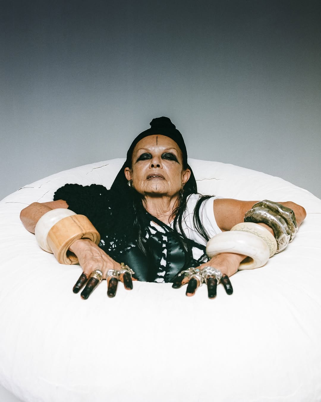 Close-up studio shot of an adult woman with dark hair, wearing statement jewelry and dark eye makeup, partially submerged in a white basin.