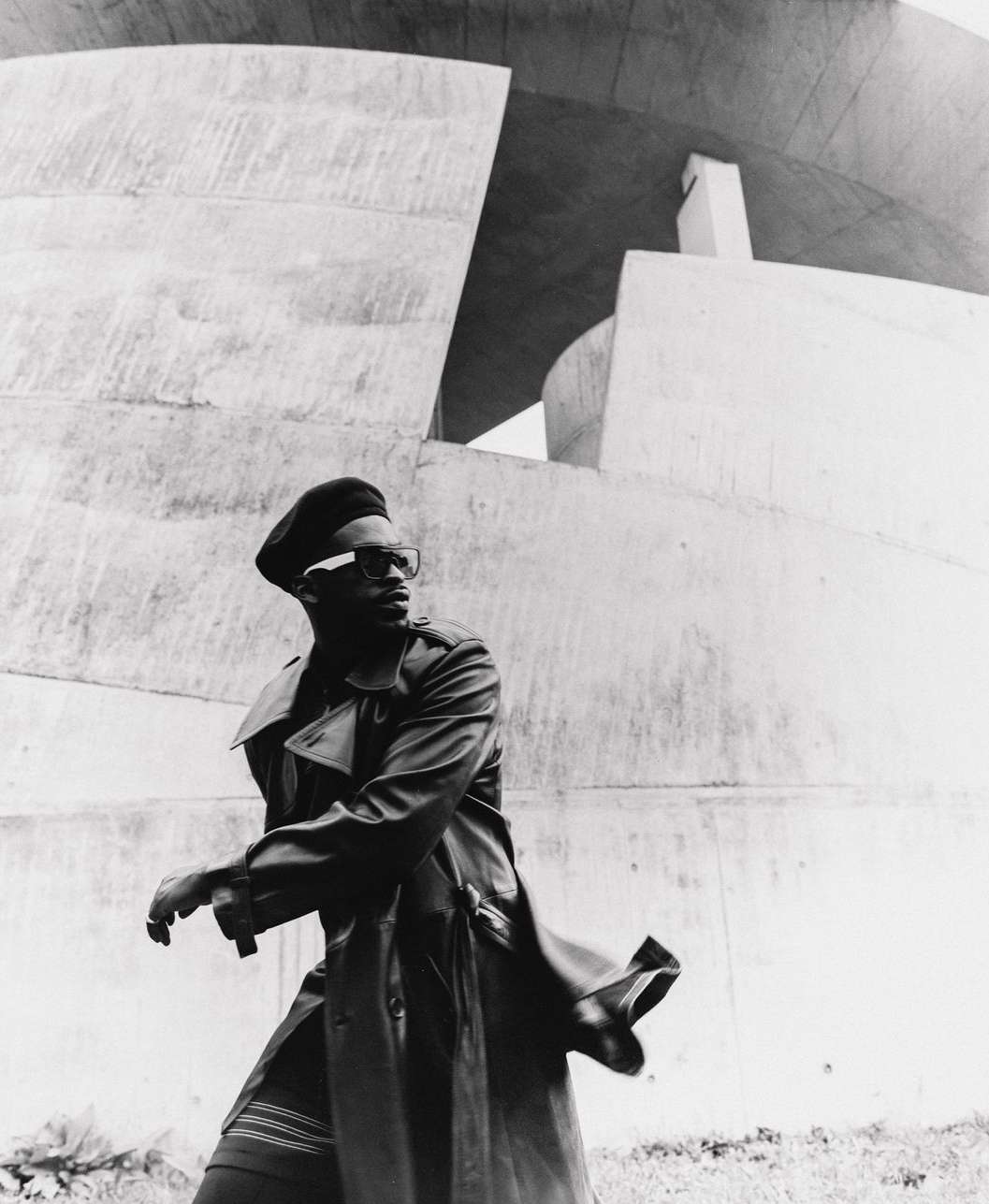 Monochrome portrait of a Black adult male in a trench coat and beret, walking in front of a concrete architectural structure outdoors.