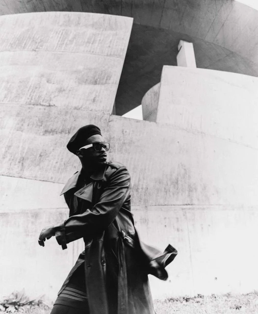 Monochrome portrait of a Black adult male in a trench coat and beret, walking in front of a concrete architectural structure outdoors.