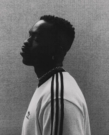 Monochrome portrait of a Black man in profile wearing a ribbed t-shirt and silver chain necklace against a textured background.
