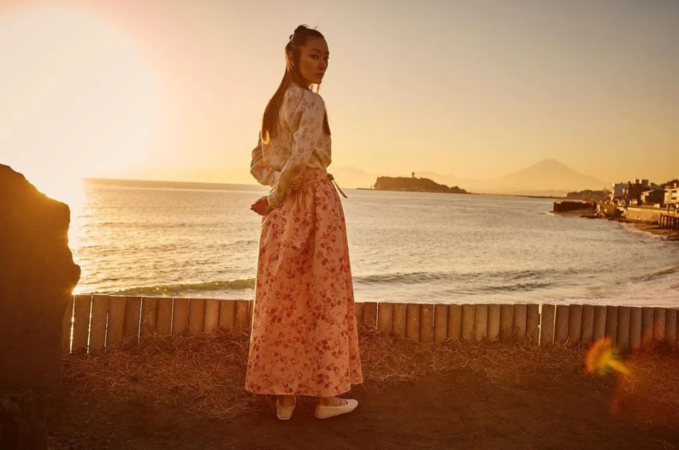 Woman in a floral skirt looking over her shoulder with a seascape background featuring an island and mountains at sunset.