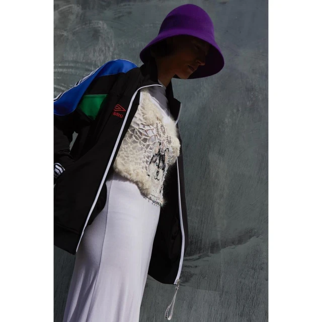 Adult female poses against textured gray wall wearing lilac dress, black jacket with blue and green accents, and purple bucket hat.