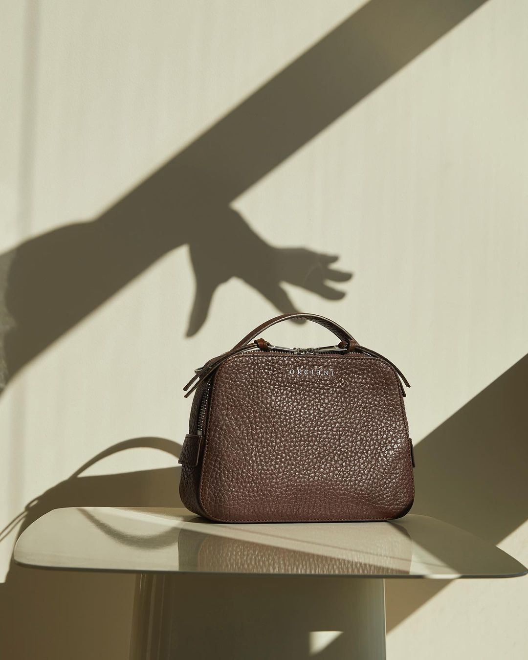Brown leather handbag on a mirrored table with a shadow hand on the wall in the background, product photography for fashion editorial.