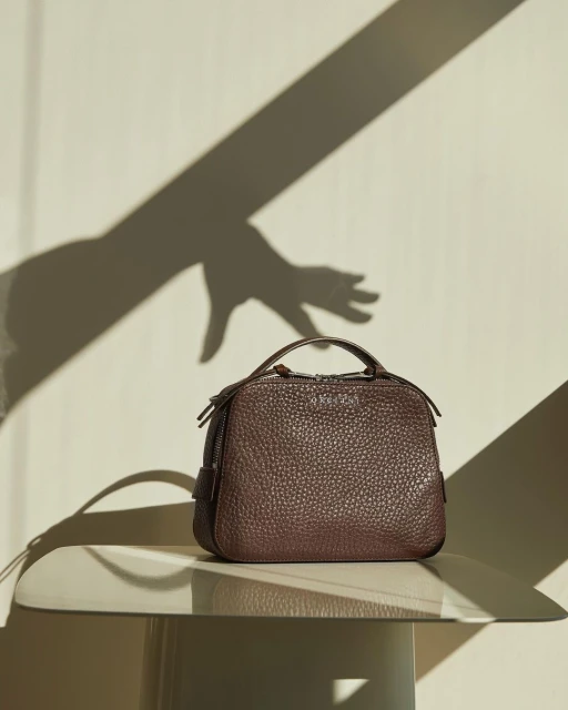 Brown leather handbag on a mirrored table with a shadow hand on the wall in the background, product photography for fashion editorial.