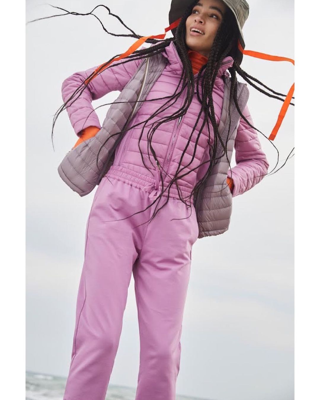 Young woman poses in a pink quilted snowsuit and grey vest, set against an outdoor backdrop with a light sky and sandy beach.