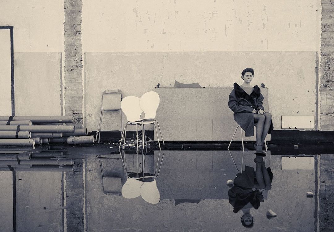 Monochrome fashion portrait of a woman sitting on a chair indoors, with a reflection in the water on the floor, creating a surreal atmosphere.