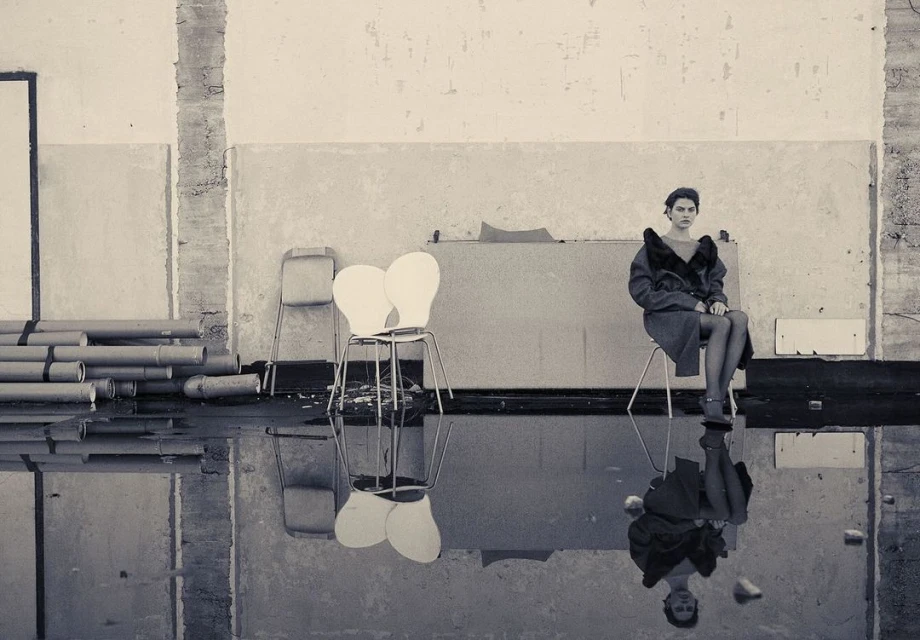 Monochrome fashion portrait of a woman sitting on a chair indoors, with a reflection in the water on the floor, creating a surreal atmosphere.