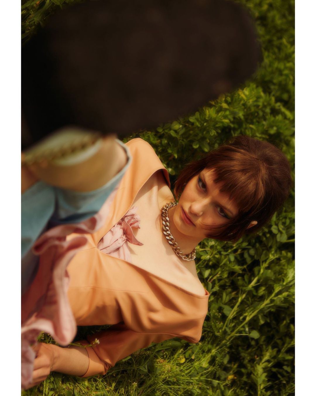 Brunette woman lies in a peach dress with a floral pattern, silver necklace, and full face of makeup, lying in the grass. 