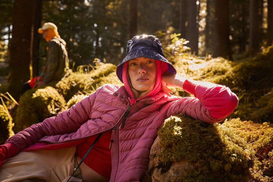 Woman in pink jacket and blue bucket hat rests on mossy ground in a sun-drenched forest, another person blurred in background.