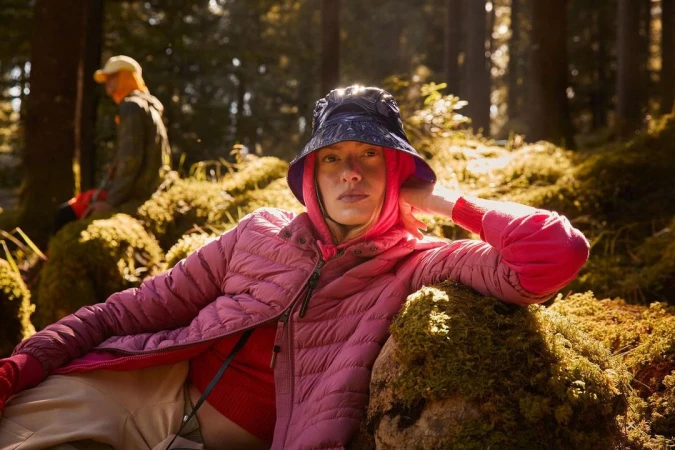 Woman in pink jacket and blue bucket hat rests on mossy ground in a sun-drenched forest, another person blurred in background.