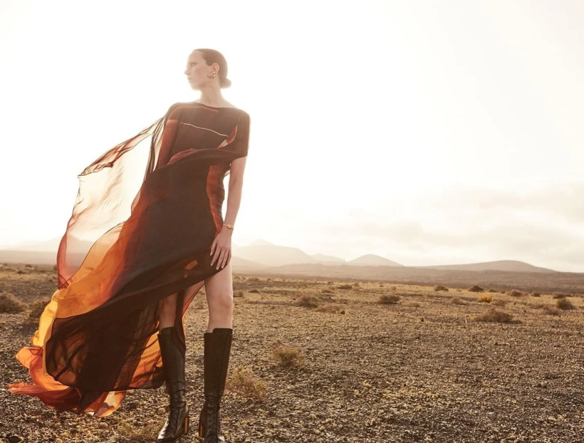 Adult female fashion model in a flowing dress and high boots poses in a desert landscape with mountains against a light sky.