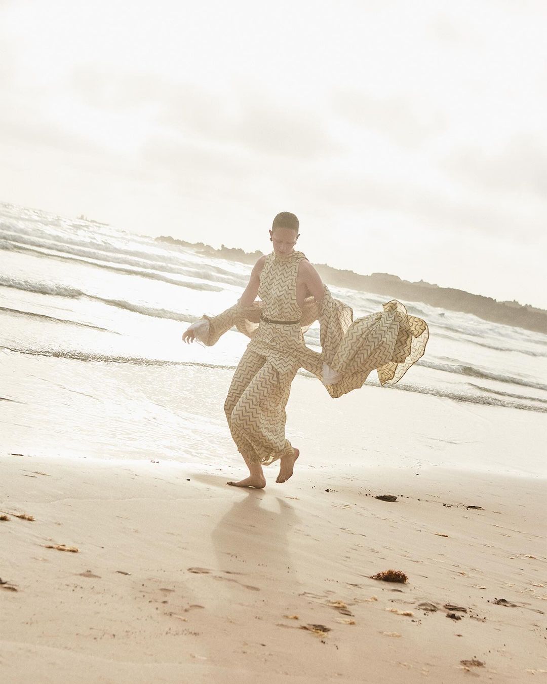 Model wearing patterned beige patterned dress walking barefoot along a sandy beach