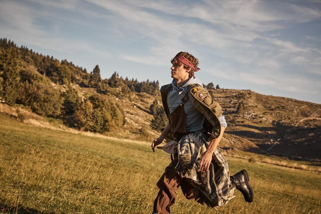A man runs through a grassy field on a sunny day, wearing a headband, hoodie, and skirt, with mountains and trees in the background.