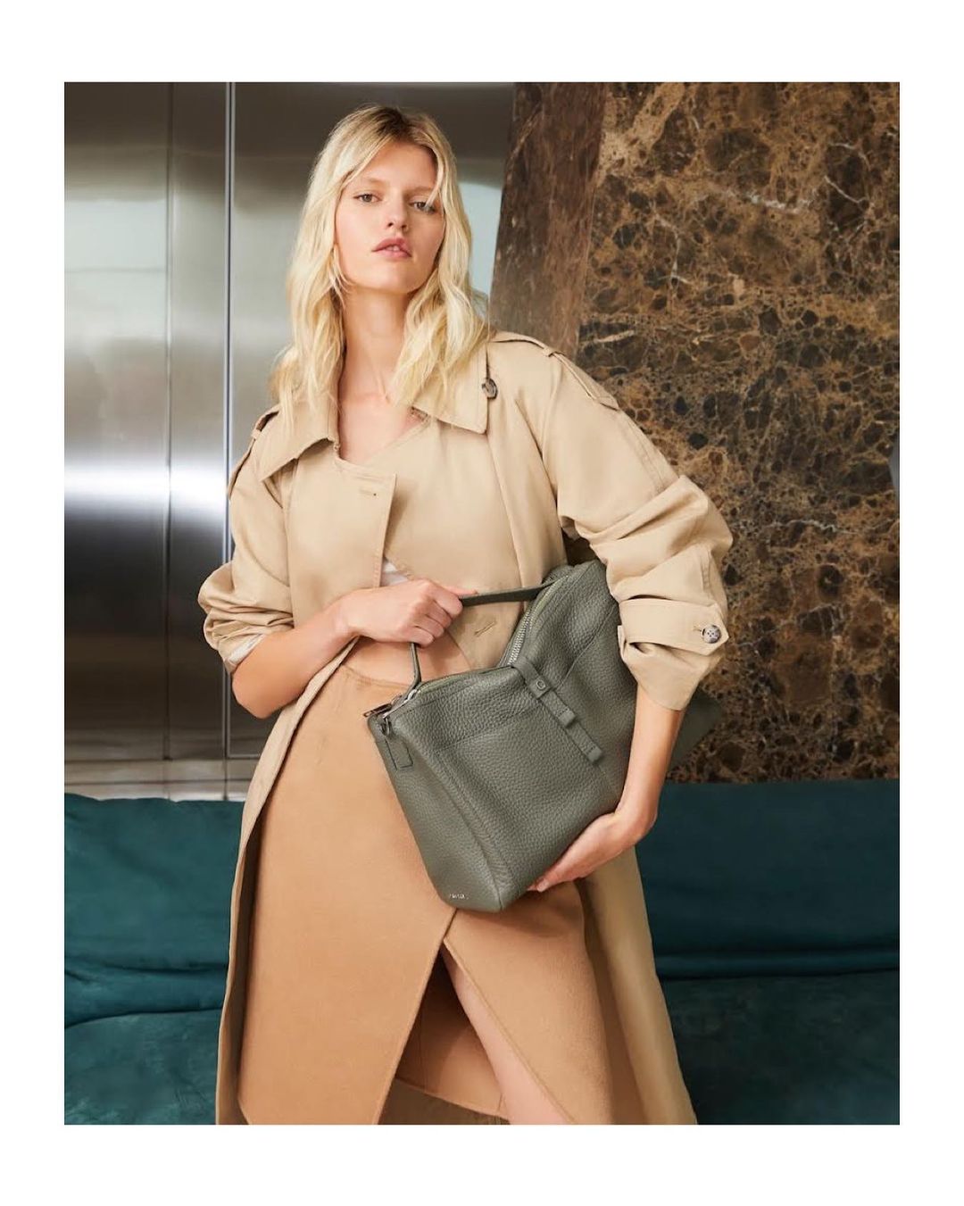 Elegant blonde model holding a textured green tote bag, wearing a tan trench coat and skirt in a stylish indoor setting with neutral tones.