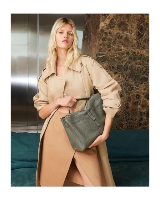 Elegant blonde model holding a textured green tote bag, wearing a tan trench coat and skirt in a stylish indoor setting with neutral tones.