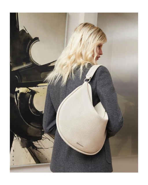 Woman in grey blazer showcasing an off-white hobo bag in a studio setting with an abstract art piece in the background.