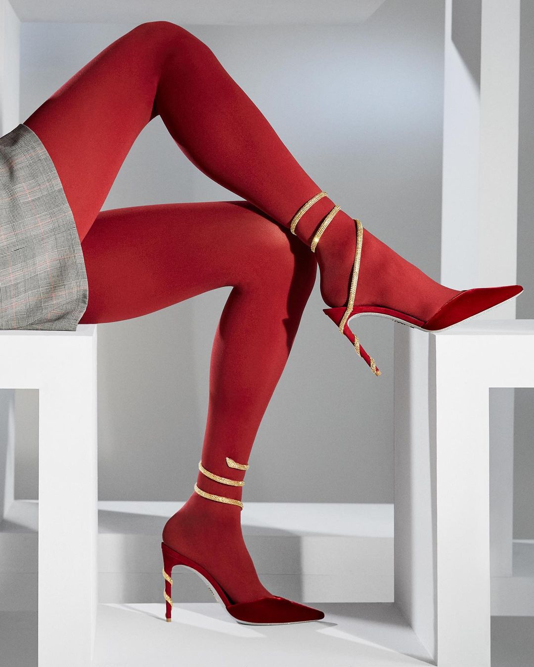 Red high heels and matching tights are presented on white geometric steps with a gray plaid jacket for a fashion editorial.