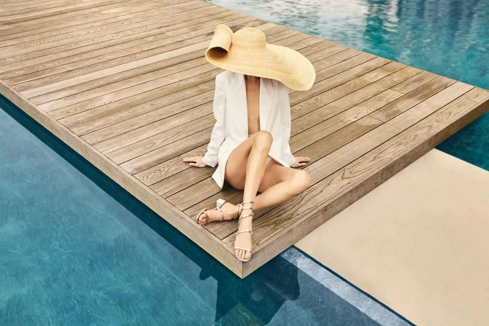 Adult female in sun hat and white jacket poses on a wooden dock next to a swimming pool in this bright, summery fashion shot.