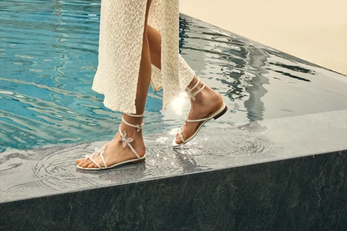 Woman in white dress stands at the edge of a pool wearing sandals with floral embellishments, highlighting fashion and luxury lifestyle.