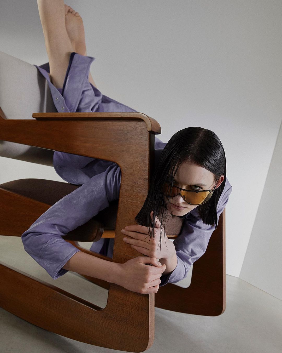 Woman in a light purple shirt and orange sunglasses reclines in a modern wooden chair in a brightly lit indoor photo studio.