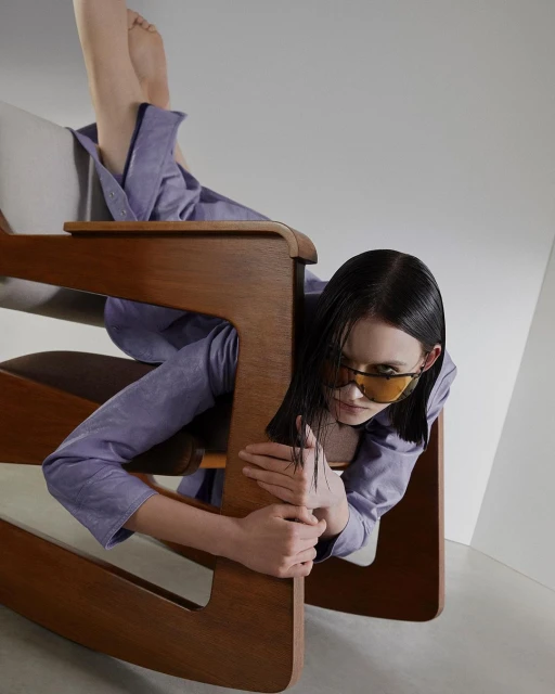Woman in a light purple shirt and orange sunglasses reclines in a modern wooden chair in a brightly lit indoor photo studio.