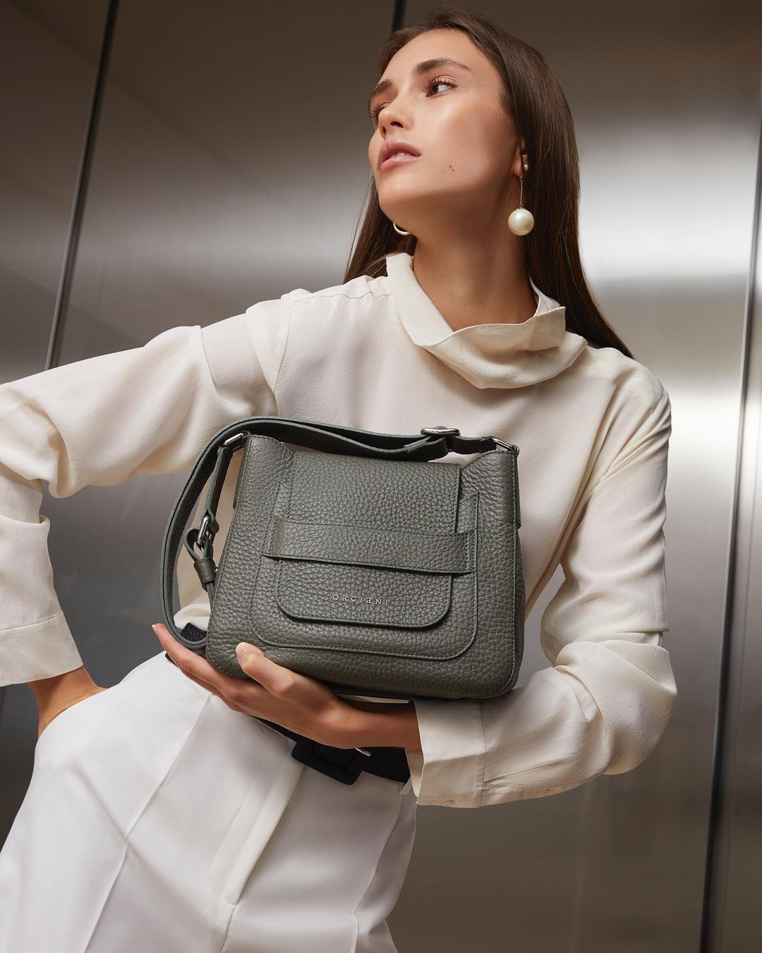 Studio shot of a woman showcasing an olive-green leather shoulder bag, accessorized with pearl earrings, against a neutral backdrop.