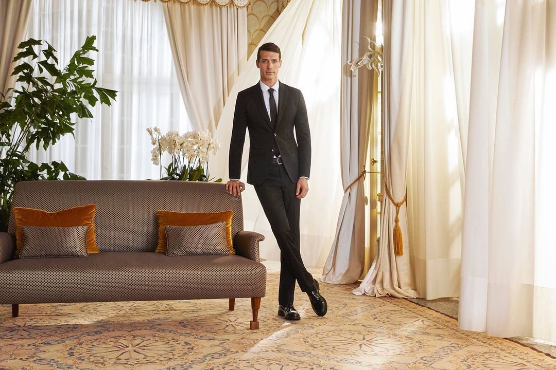 Stylish adult man in a black suit stands next to an ornate sofa with decorative pillows in an opulent indoor setting with draped windows.