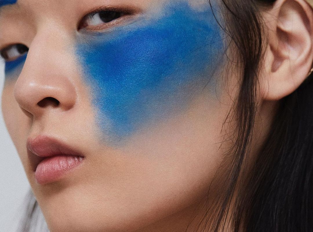 Close-up of a young woman's face with bright blue artistic face makeup. The focus is on the artistic makeup and her expression.