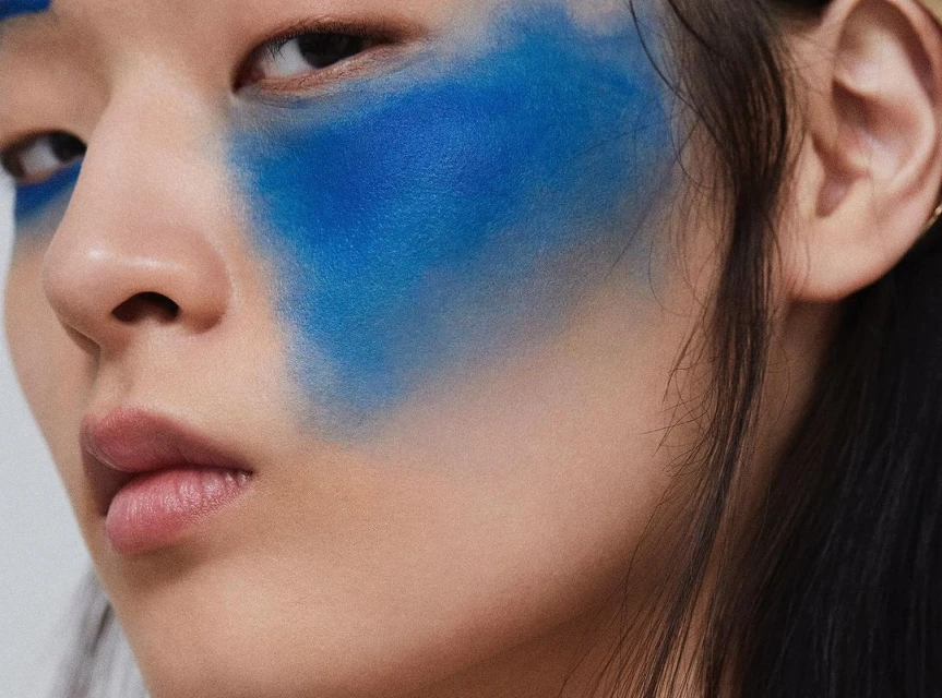Close-up of a young woman's face with bright blue artistic face makeup. The focus is on the artistic makeup and her expression.