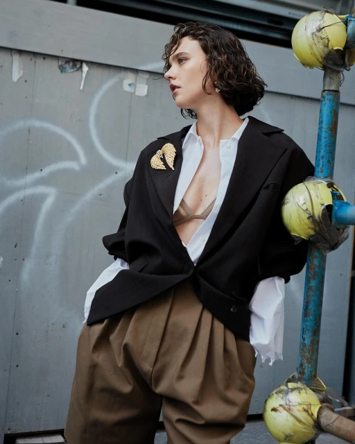 Woman in oversized black blazer with gold brooch posing in an urban setting with textured wall and industrial metal structure.
